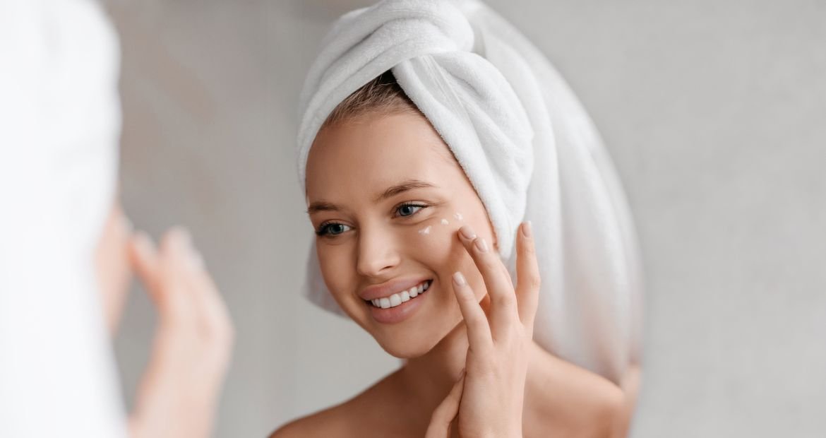Mulher sorridente com toalha branca no cabelo passando creme para olheiras em seu rosto em fundo branco.